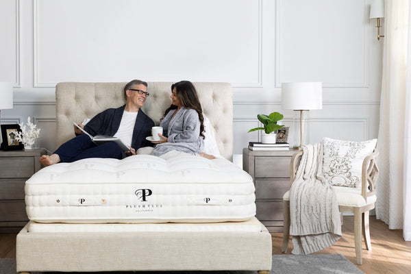 A plush mattress with a white quilted surface and the PlushBeds logo, on a bed with a woman lying and a man sitting, in a bedroom.